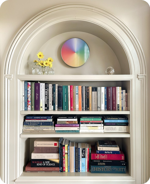 The Present annual wall clock in glass and steel, front view, displayed above a bookshelf in a home interior.